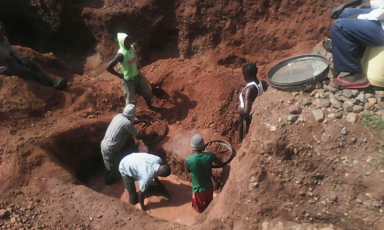 A team of six miners use sieves to sift through red mud in search of diamonds, while a supervisor looks on from the mine’s rim.