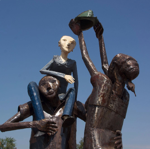 Cast against a clear blue sky, a metal sculpture depicts three people: a light skinned child atop their dark skinned father’s shoulders, while their dark skinned mother reaches out with a protective sun hat.