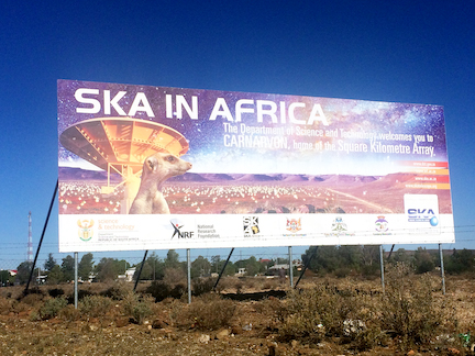 The image shows a huge billboard. On the left is a meerkat, behind it is a telescope dish. In the background one sees many more dishes on a plain in front of some mountains in the distance. Against a starry sky, the billboard reads: “SKA in Africa, The Department of Science and Technology welcomes you to Carnarvon, home of the Square Kilometre Array.”
