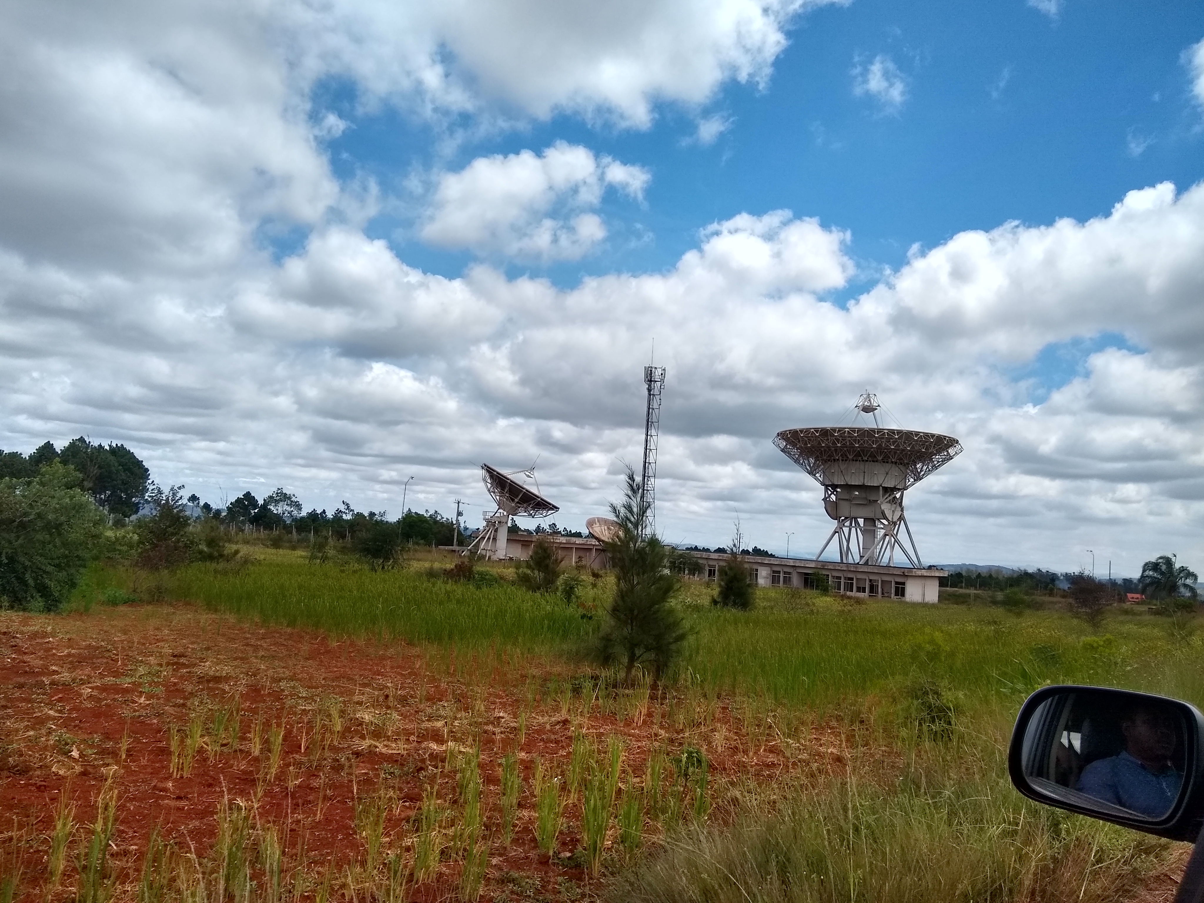 Central to this image is a gigantic satellite dish with a smaller dish that is slightly tilted. At the foot of the big dish is a single-story house. In the foreground is a patch of land that was used for agriculture.