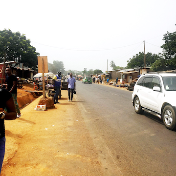 On a clear day, a man wearing a blue shirt and slacks walks along the highway lined with various vendors at a security checkpoint to wait for the bus to pick up passengers as a white car drives past.