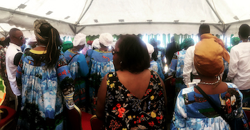 Rows of men and women stand below a white tent wearing the thematic print for the Ngondo Festival, featuring a mostly blue with images of boats, trees, and clouds. In the center of the photo stands an African American woman wearing sunglasses wearing a Black floral print dress.