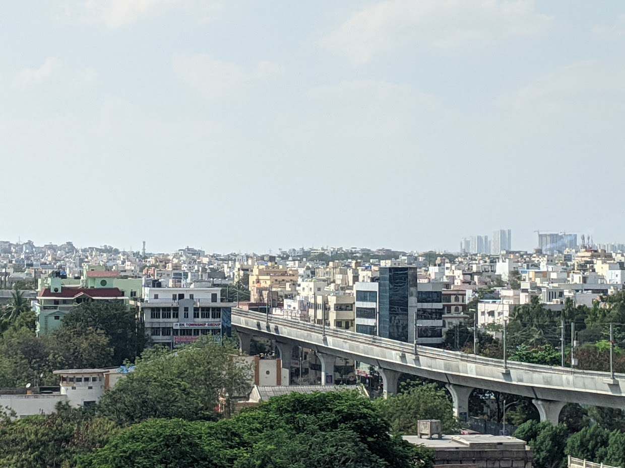A skyline photograph of Hyderabad, showing trees and a metrorail track in the immediate foreground, with sprawling built environment stretching to the horizon. In the distance, there are high-rise towers under construction.