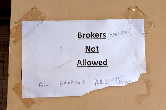 A white poster on a wall printed with the words “Brokers Not Allowed”. There are handwritten vandal annotations naming a particular broker, and a comment saying “All brokers are th… [obscured by tearing]”.