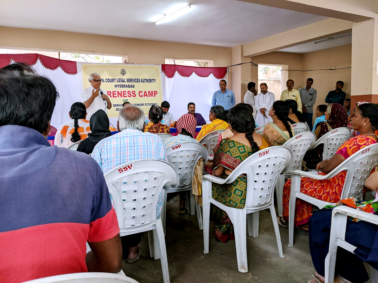 Photograph of a public meeting where a grey haired man is addressing an audience seated on plastic chairs. Next to the speaker are other panelists seated at the front of the meeting space. There are also a group of men standing to the side. Behind the speaker, there is a banner that reads “City Civil Court Legal Services Authority, Hyderabad. Awareness Camp”.