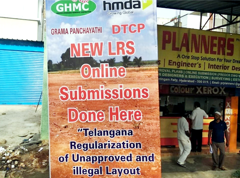A freestanding banner announcing a new layout regularization scheme. The banner reads “New LRS: Online Submissions Done Here. Telangana Regularization of Unapproved and illegal Layout Scheme.” The banner includes logos of the Greater Hyderabad Municipal Corporation, the Hyderabad Metropolitan Development Authority, the Department of Town and City Planning, and the Department of Gram Panchayati (rural governance). In the background is a storefront belonging to “planners”.