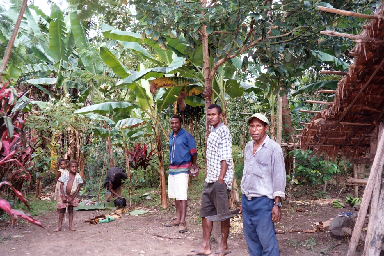 Image shows three men, two children and a woman bent over a fire in the middle of their coffee grove preparing to work. One older man wearing a baseball cap carries a machete while standing next to a house made of bush materials.
