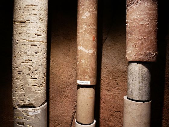 Several cylinders of rock, each of a different composition, hung vertically in three rows against a wall.