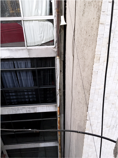 A shadowy gap sits between the wall of a building and an external retaining wall, which seems to be widening toward the top. On the left, the curtains and bedding of a bedroom are visible.