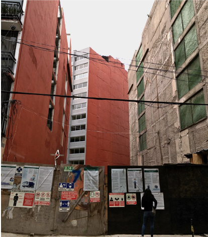 A boarded up vacant space between two buildings. A person stands in the foreground looking at the emptiness.
