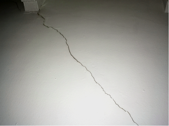 White wall with a wooden beam at the top, and a large crack snaking diagonally downward to the right.