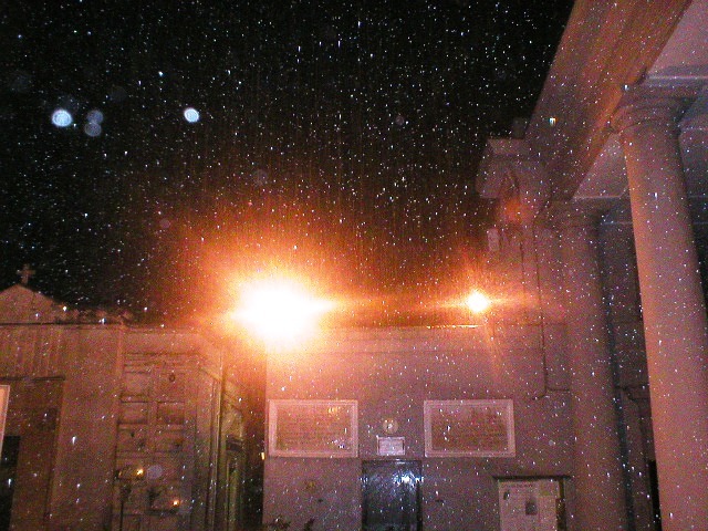 Nighttime view of a building courtyard with columns on the right and a low wall in the background. Two bright lights flare toward the camera, illuminating falling dust against a dark sky.