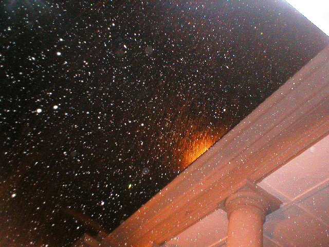 Nighttime view of a courtyard with columns on the right and a low wall in the background. Two bright lights flare toward the camera, illuminating visible dust particles suspended in the air against a dark sky.