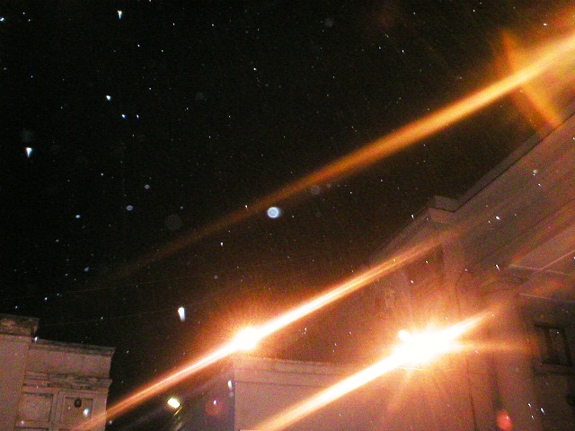 Nighttime view of a courtyard with classical architecture. Two bright lights create diagonal flares across the image, illuminating visible dust particles suspended in the dark sky.