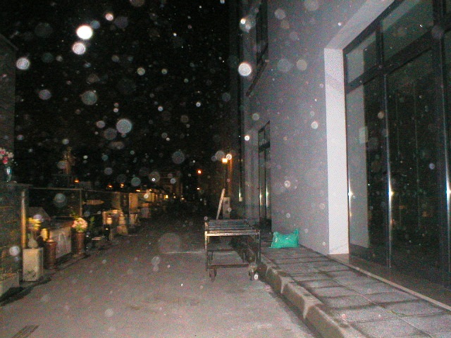 Nighttime street beside a building, with visible dust illuminated in the air. A bench and doorway line the right side; graves and small lights recede into the distance on the left.