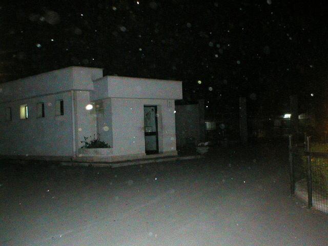 At night, a small white building stands along a paved path, with visible dust illuminated in the air against a dark sky.