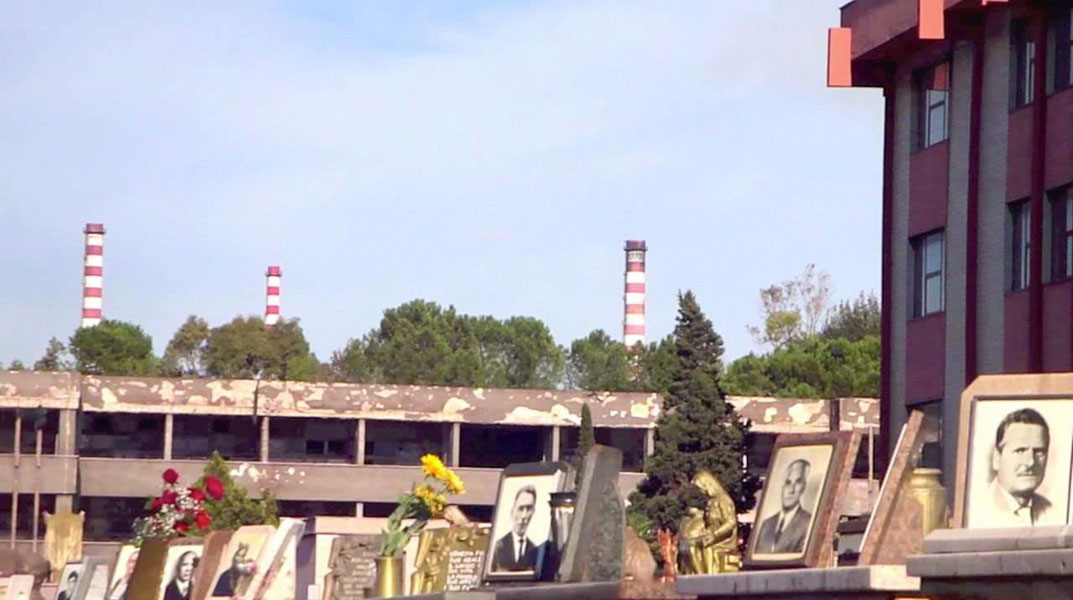 Daytime view of a cemetery with grave markers and framed portraits in the foreground. In the background, an industrial building and striped smokestacks rise behind trees.