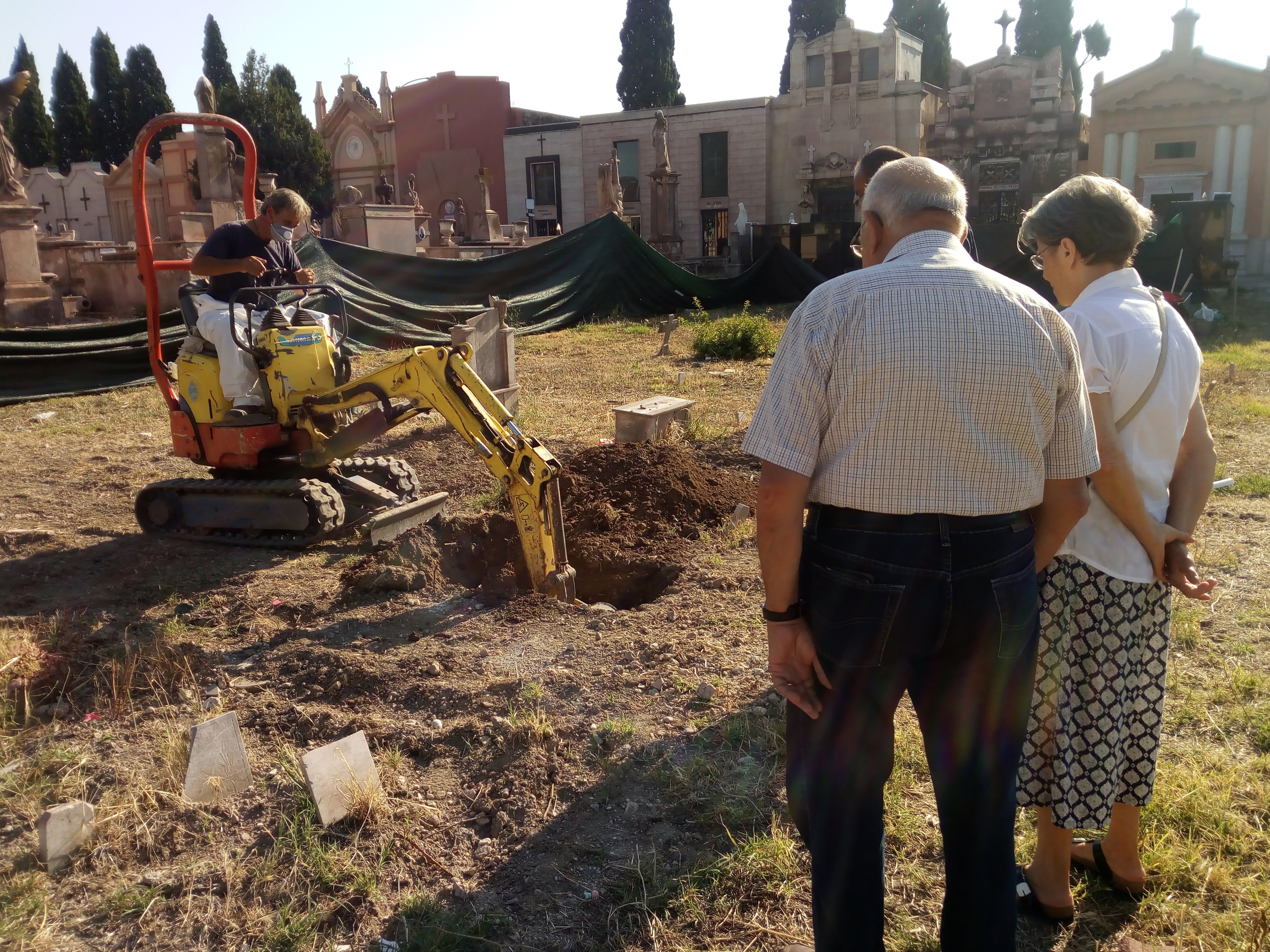 In a cemetery during the day, a small excavator digs a hole in the ground while two people stand nearby watching. Tombs and mausoleums line the background.