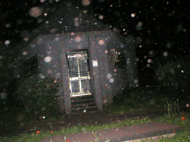 At night, visible dust particles fill the foreground in front of a small building with a lit doorway and steps, surrounded by grass.