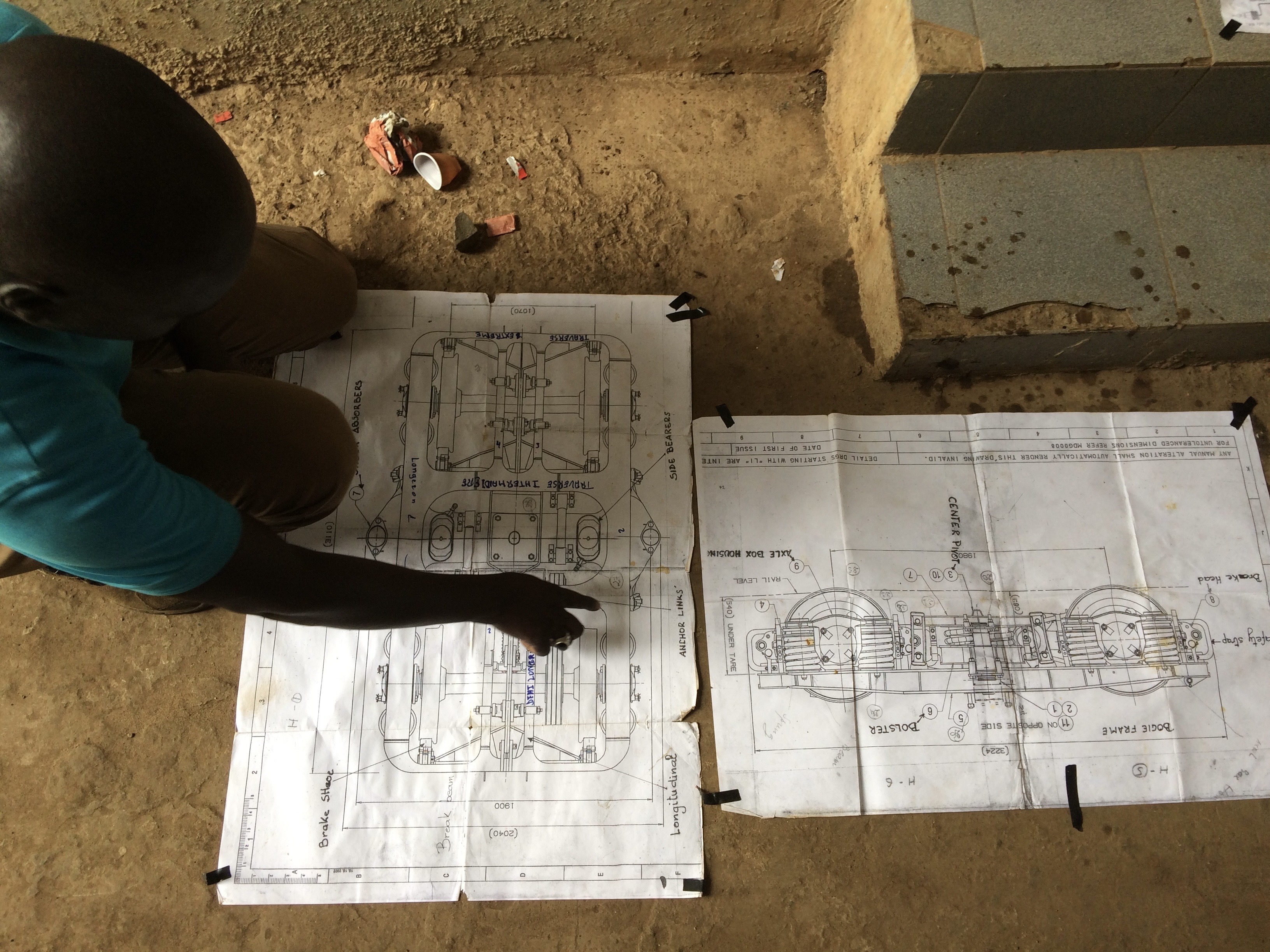 A man crouches on the ground in Thiès, Senegal, pointing at technical blueprints of train axles and bogies spread out on the floor, annotated with French translations.