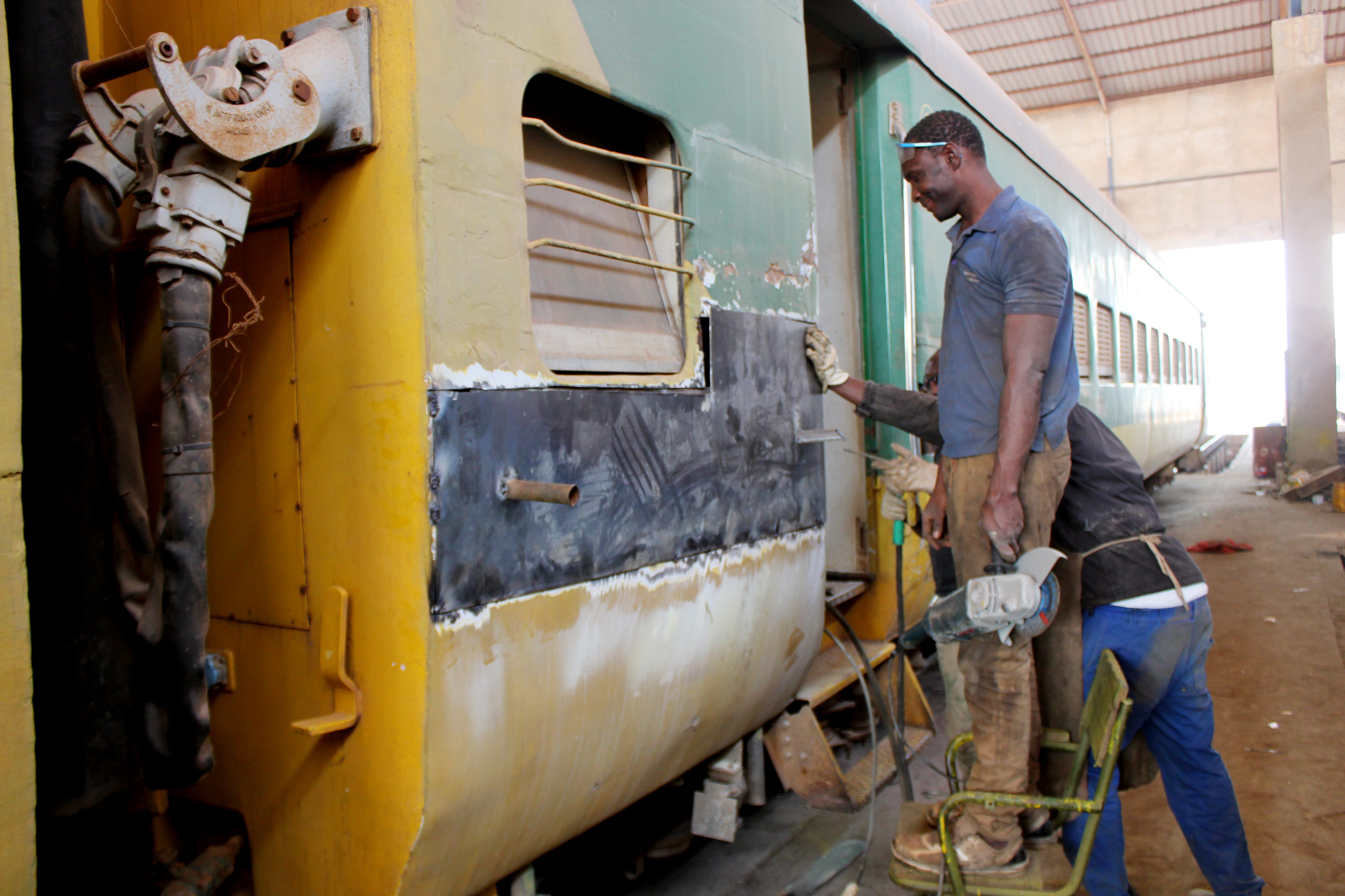 Two railway workers patching the side of a yellow and green passenger carriage inside a workshop, one holding a grinder while the other welds a new metal sheet onto the body.