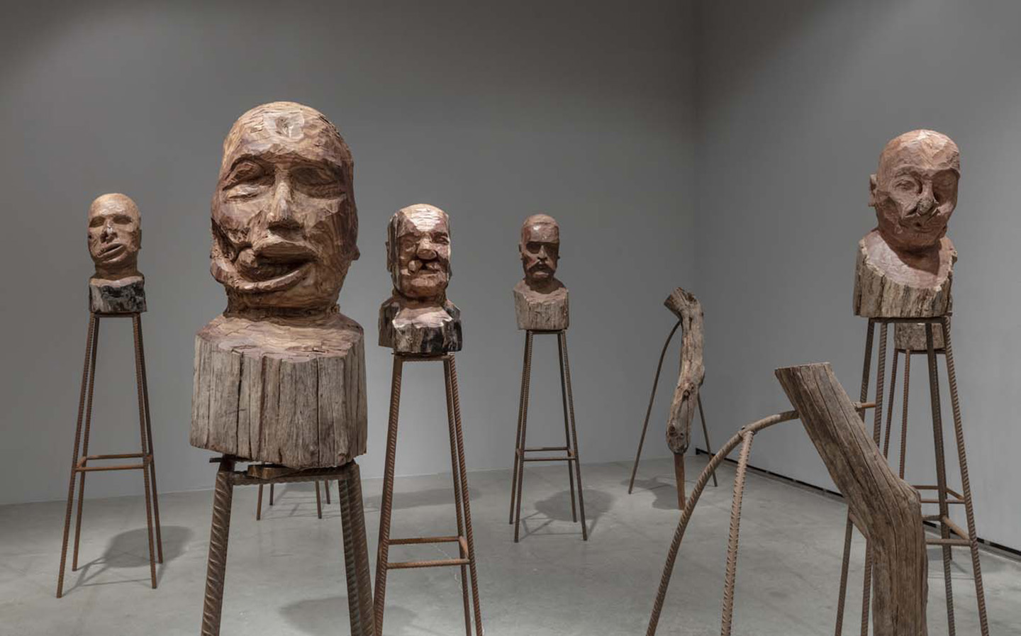 Installation view of ‘J’accuse,’ showing six wooden busts with rough-carved human faces, created by Kader Attia with Senegalese craftsmen, mounted on tall metal supports in a gallery space.