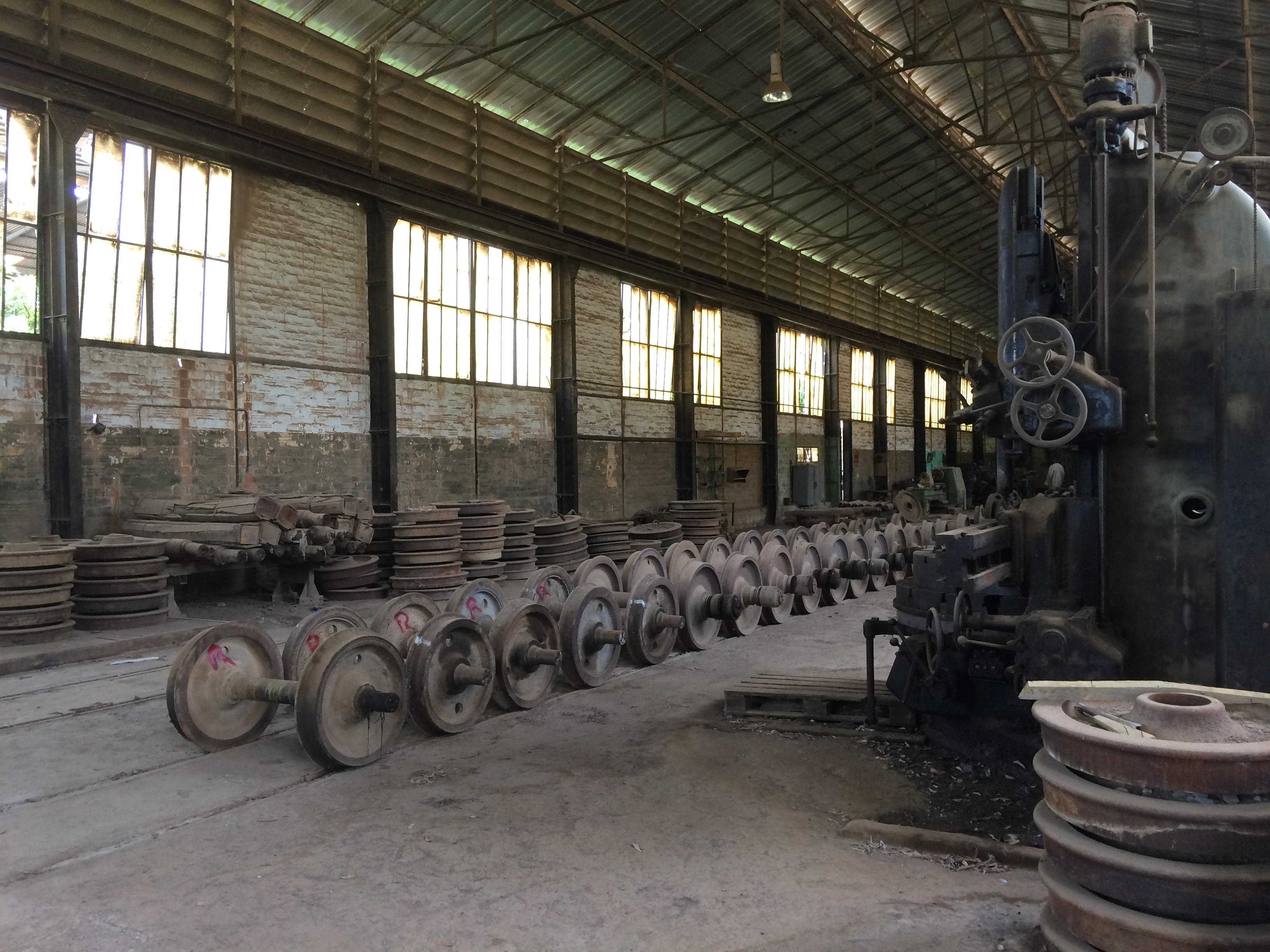 Rows of dismounted train axles and wheels lined up inside the Thiès railway workshop in Senegal, with stacks of spare parts and a large industrial machine visible in the background.