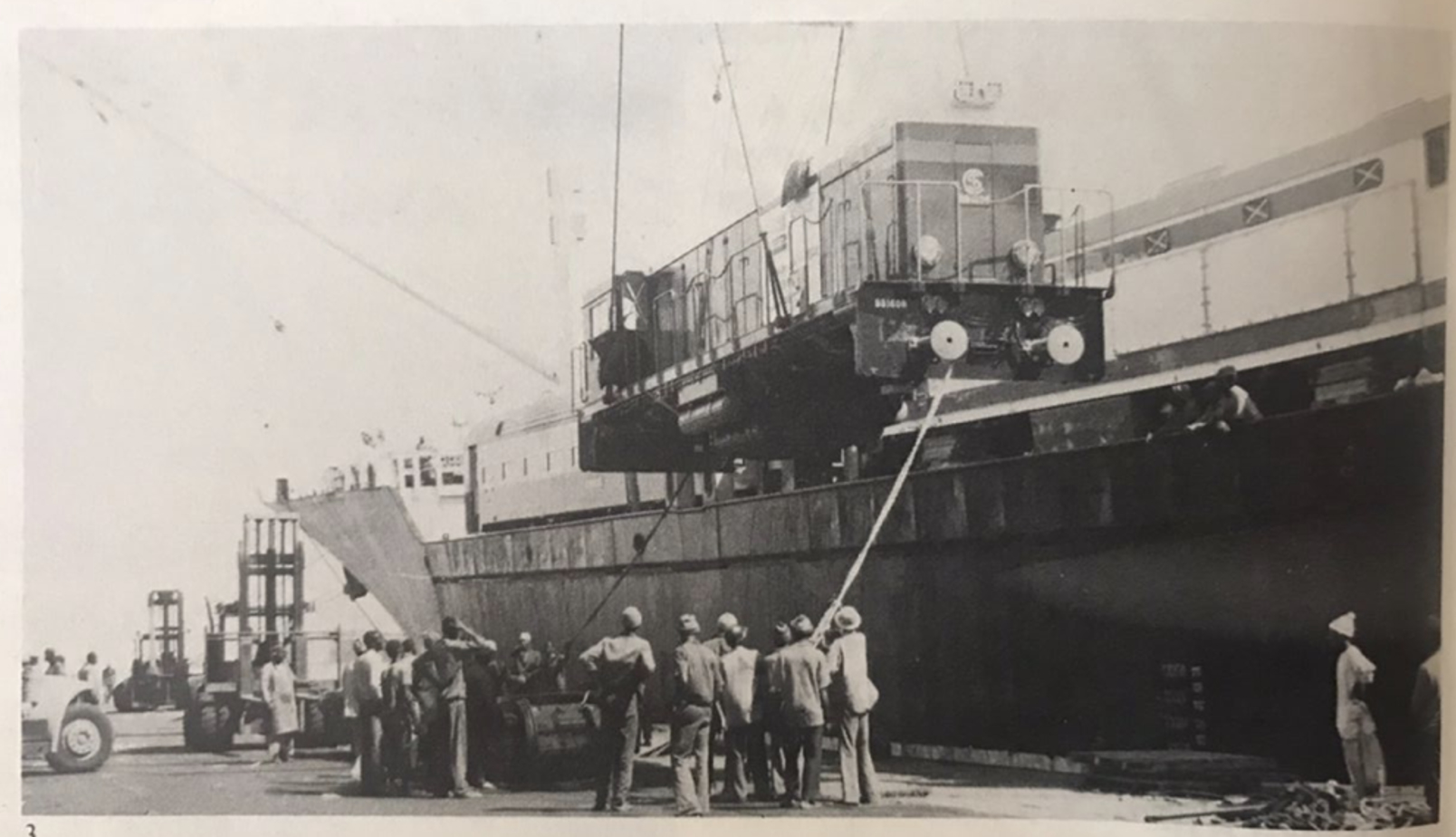 Black-and-white photograph of a locomotive being lifted by crane onto a ship, with dockworkers gathered below watching the transfer.