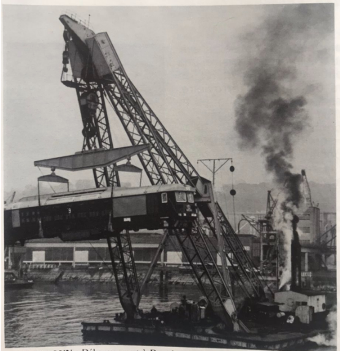 Black-and-white photograph of a large crane hoisting a passenger train carriage onto a ship at a port, with smoke billowing from the crane’s steam engine.