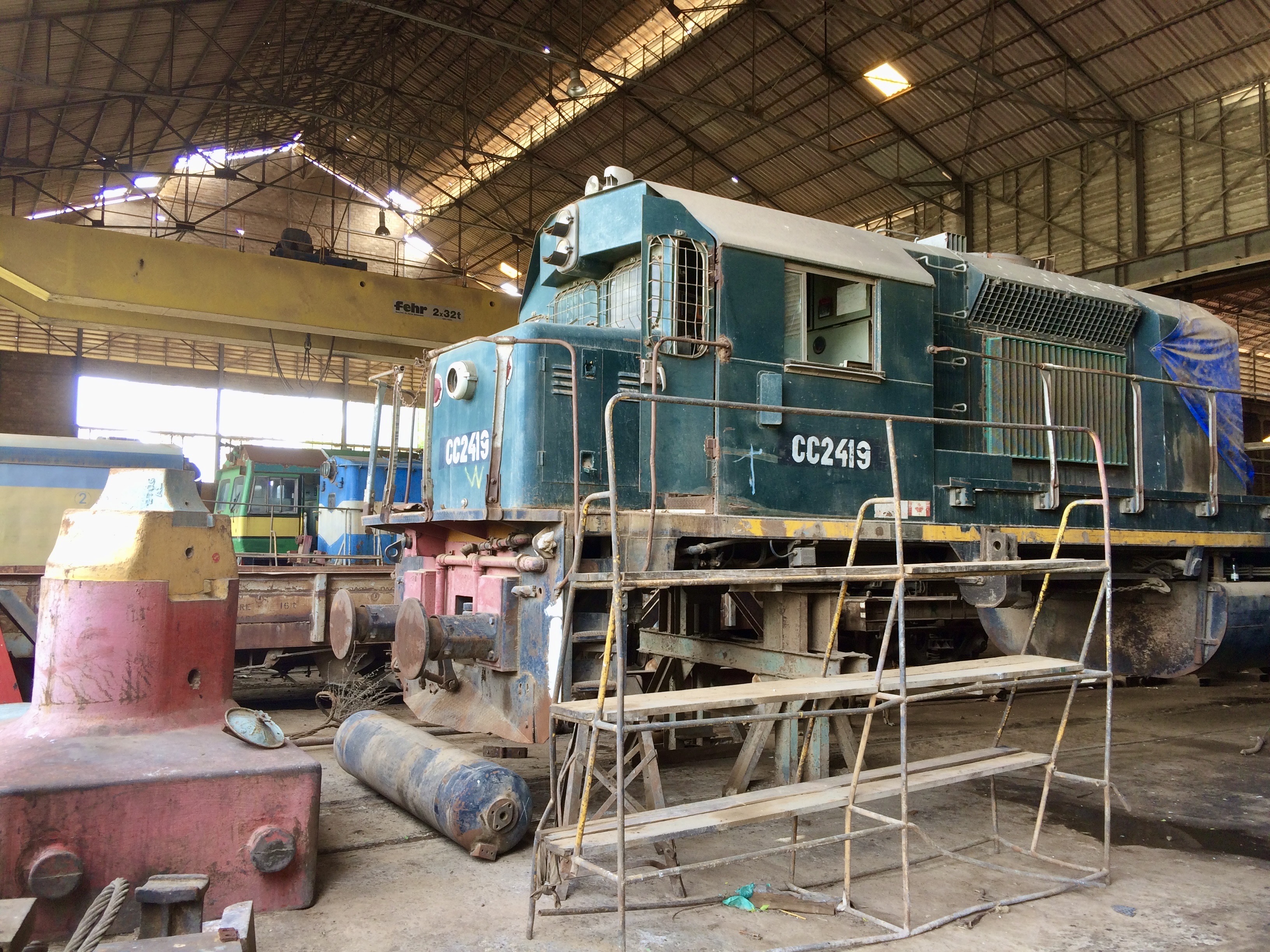 Green diesel locomotive CC2419 under repair inside the Thiès railway workshop, elevated on supports with scaffolding in front and other rail equipment visible in the background.