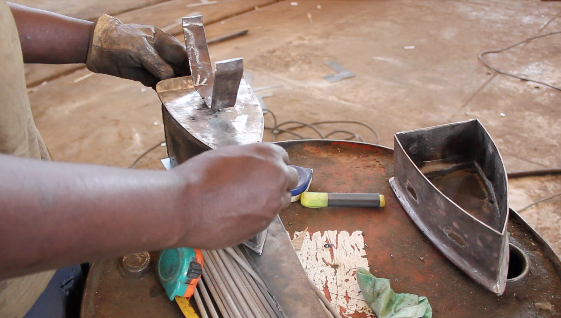 In the Rufisque workshop, a railway worker shapes surplus sheet metal into a household iron, welding the handle onto the base.