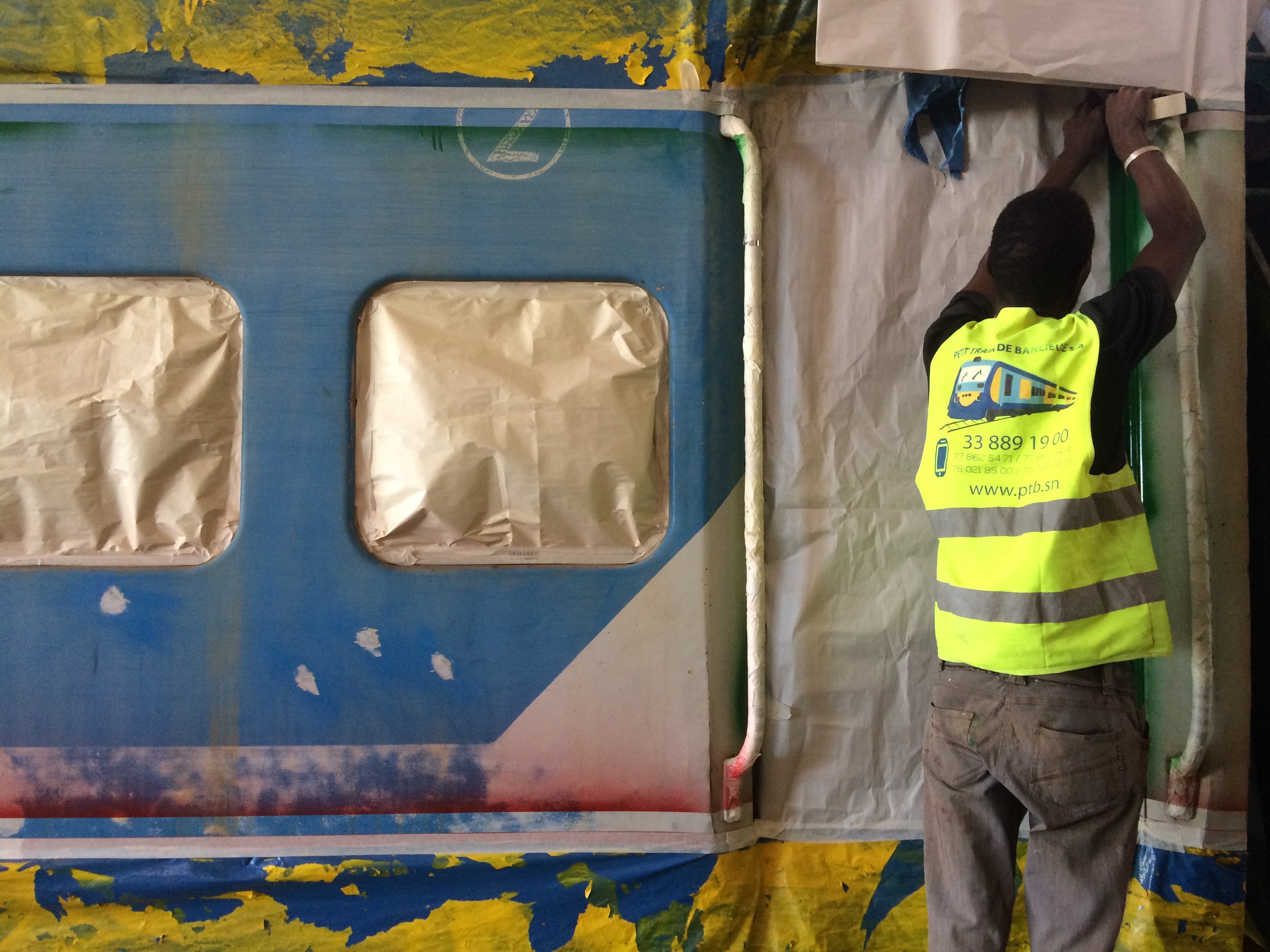 Railway worker in a reflective vest masking the windows and doors of a blue train carriage with paper and tape in preparation for repainting.