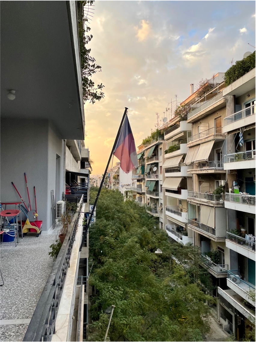 Pastel flag hanging off a fourth floor balcony with a tree lined street below.