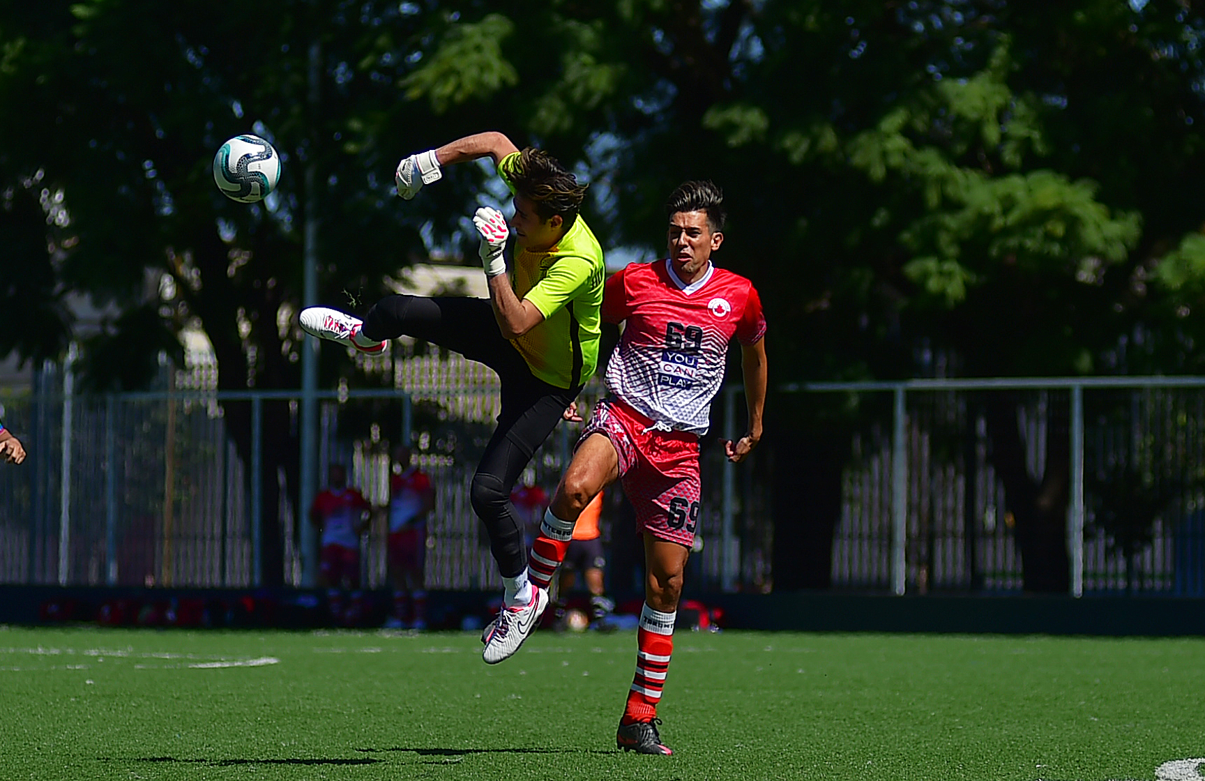 A football goalkeeper jumps and kicks the ball in the air while under pressure from an opponent.