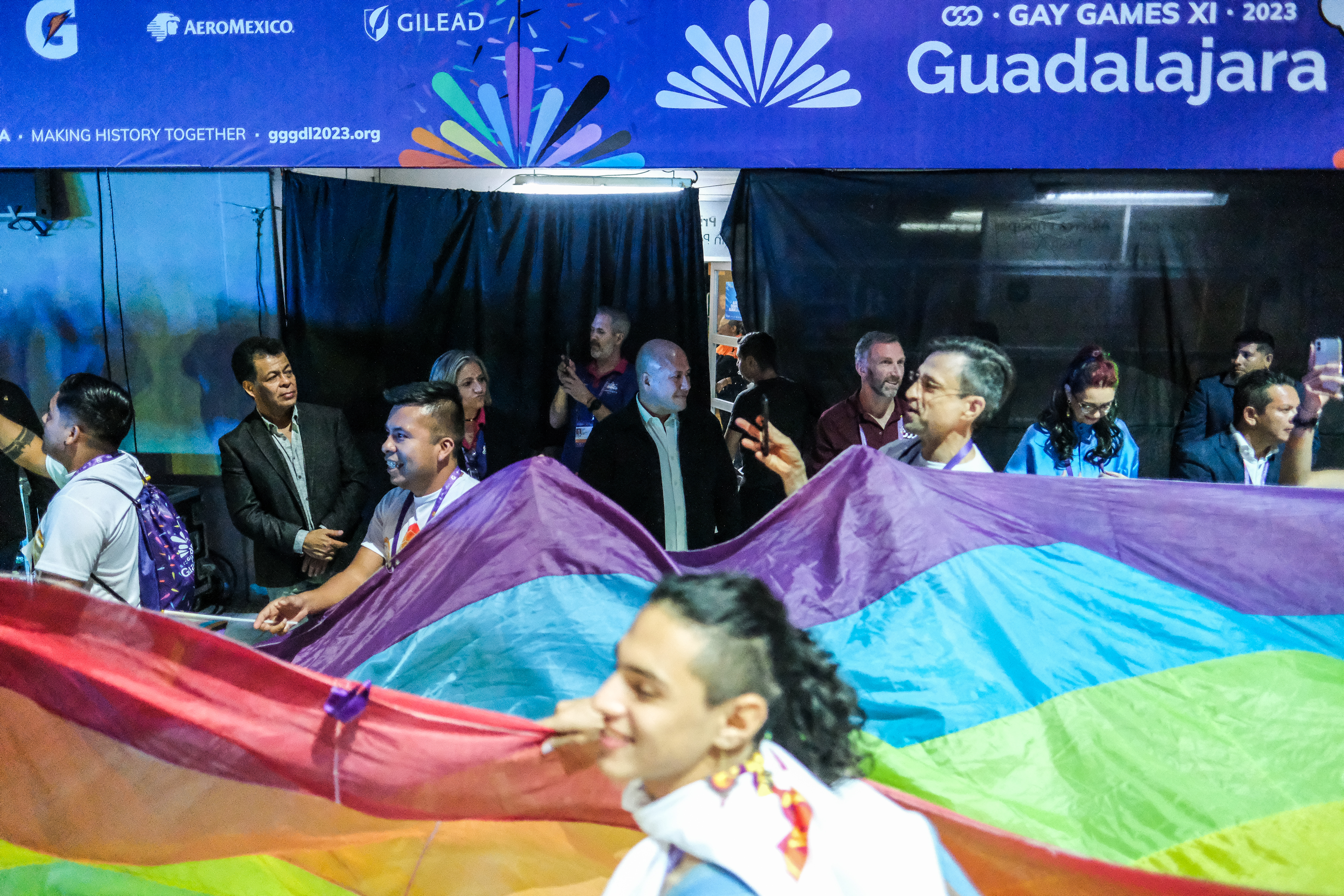 Athletes carry large rainbow flag in an indoors venue with politicians behind them.